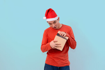 young man with sober expression in santa hat holding holiday gifts against blue background