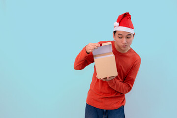 happy young man dressed in a festive outfit, wearing a Santa hat, holding an open gift box for christmas concept against blue background