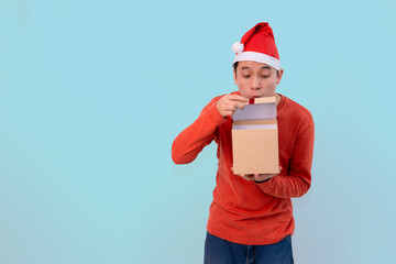 happy young man dressed in a festive outfit, wearing a Santa hat, holding an open gift box for christmas concept against blue background