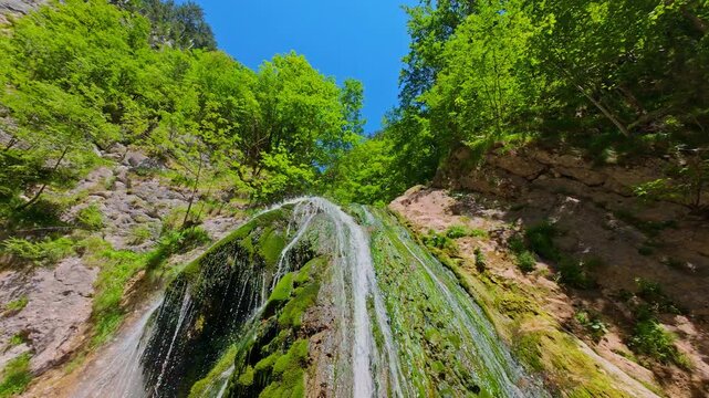 Aerial video of a scenic waterfall cascading down a rocky mountain cliff surrounded by lush green trees and forest, captured in smooth FPV drone slow motion on a sunny summer day.