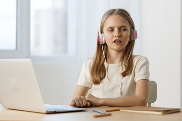 A thoughtful girl wearing headphones sits at a desk with a laptop, focused on her work, in a bright and modern workspace