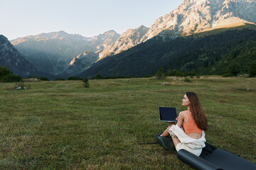 Fototapeta premium A woman sits on a grassy meadow with a laptop in a remote mountain setting. She appears focused and calm, enjoying a peaceful, productive outdoor workspace with expansive scenery and blue sky above.