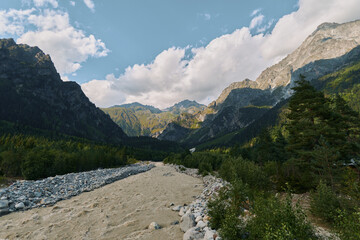 A pristine mountain valley unfolds with a winding river, rocky banks, and evergreen forest under a bright sky, creating a tranquil outdoor scene rich with natural beauty and expansive scenery.