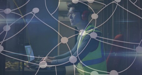 Technician wearing vest, headphones using laptop at data center, with server racks, network cables