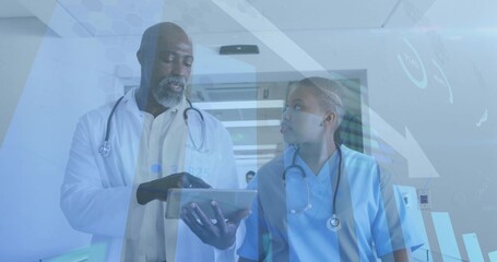 Walking lab coat doctor and scrubs nurse with stethoscopes reviewing tablet in hospital corridor