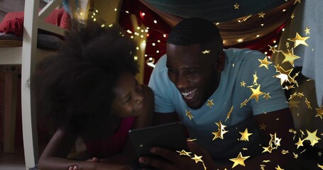 Lying father and daughter reading tablet inside draped sheet fort at home, with string lights