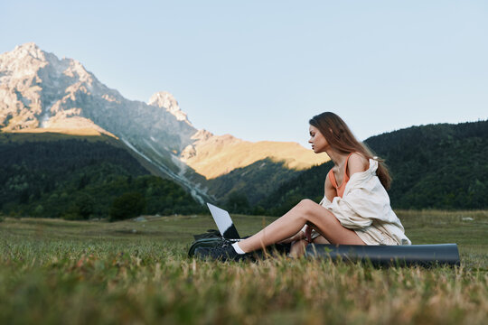 A woman sits on a blanket in a vast meadow, typing on a laptop outdoors with mountains in the background, conveying focus, creativity, and remote work in nature.