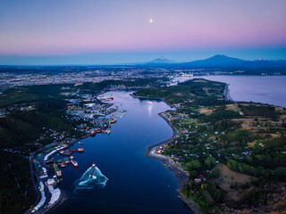An aerial view of Puerto Montt's Canal de Tenglo and Tenglo Island during the blue hour, with a full moon reflecting on the water and the Osorno volcano in the distance.