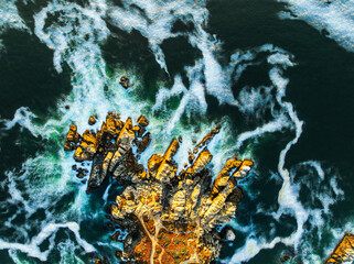 An abstract aerial long exposure of a rugged Pacific coastline, with golden-lit rocks surrounded by silky, white turbulent water at sunset