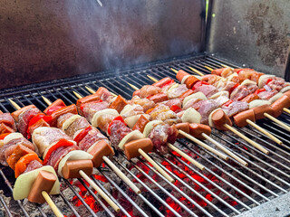 A close-up, low-angle view shows traditional Chilean anticuchos - skewers of meat, sausage, and vegetables - sizzling on a hot charcoal grill with a vibrant glow from the coals below.