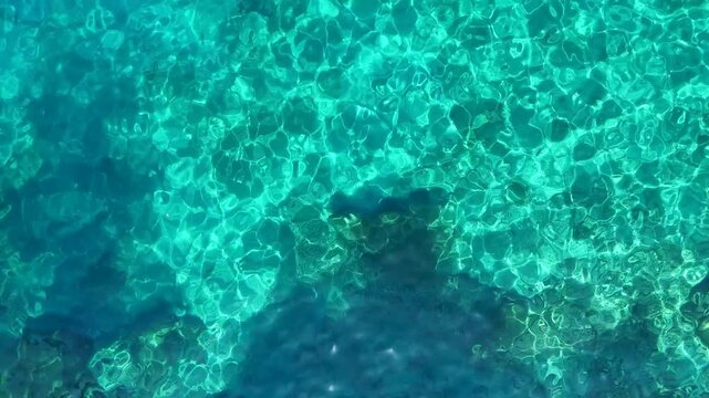 An abstract aerial video of crystal clear turquoise water with the rocky sea bed visible below creating a beautiful and mesmerizing natural pattern with light reflections