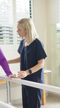 Female therapist guiding female patient practicing walking in rehab with parallel bars