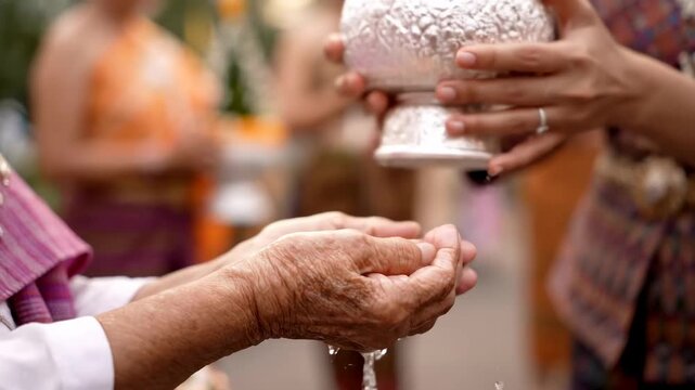 Close up pouring water elderly hands blessing cinematic UHD handheld Songkran tradition lifestyle stock. ablution aged palm benediction liquid cascade thai new year custom