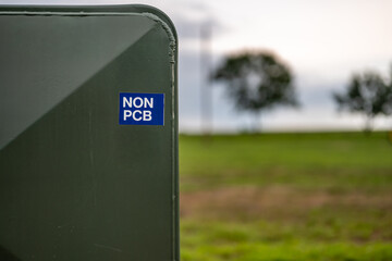 Utility box marked as non-PCB located in a green field with trees in the background during overcast...