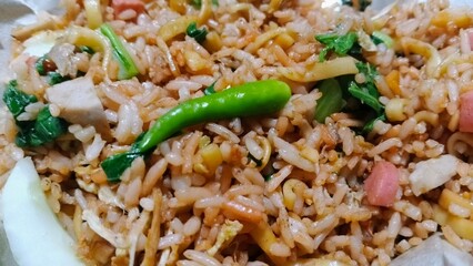 Macro photograph of Indonesian fried rice (Nasi Goreng). A close-up view showing rice texture, a green chili, chopped vegetables, sausage, and egg.