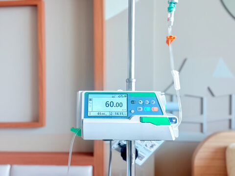 Medical infusion pump with digital screen displaying flow rate in a hospital room. Pediatric patient resting in bed in the background. Healthcare and intravenous therapy concept.