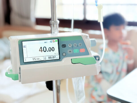 Medical infusion pump with digital screen displaying flow rate in a hospital room. Pediatric patient resting in bed in the background. Healthcare and intravenous therapy concept.