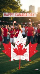 A large, decorative maple leaf, adorned with Canadian flags and fireworks, stands in a grassy area at a public celebration.