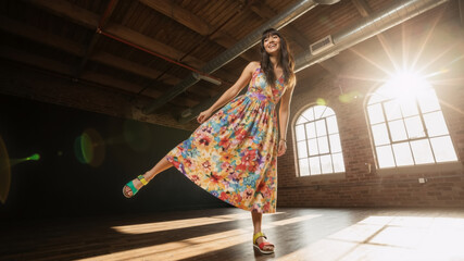 Happy woman in a colorful floral dress dancing playfully in a sunlit industrial loft. Summer fashion and lifestyle portrait. Backlit with lens flare.