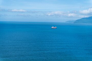a lone cargo ship sailing across the vast, deep blue sea near Hai Van pass, with distant coastal mountains visible under a cloudy sky.