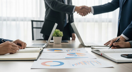 Business Collaboration and Data Analysis. Charts and notebooks are laid out on the conference table, while two people shake hands in the background,  conveying an atmosphere of cooperation
