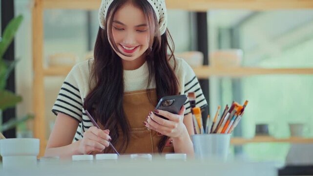 Happy Asian female artist using smartphone to find references and ideas for painting ceramic pottery design in creative studio, woman painting custom artwork for client project