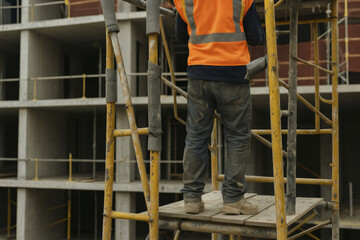 Construction worker stands on scaffold, wearing safety vest and sturdy footwear. background features building under construction, highlighting importance of safety in construction environments