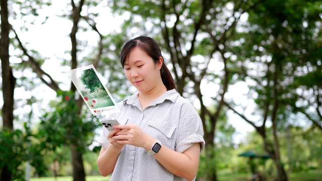 Happy young Asian woman using a smartphone, checking social media notifications with likes and heart icons on her photo post, enjoying a relaxing day in the park.