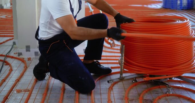 Unrecognizable worker unrolling coil of plastic pipes for underfloor heating installation indoors, preparing floor heating system during construction.