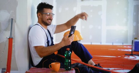 Professional construction worker sitting on floor indoors during underfloor heating installation break, laughing while having video call on smartphone.