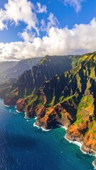 Verdant, jagged coastal cliffs meet vibrant turquoise ocean under a partly cloudy blue sky in a stunning aerial perspective