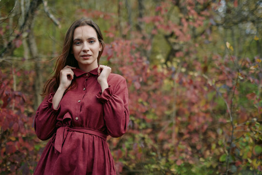 A candid portrait of a woman in a burgundy dress, conveying authenticity and credible presence amid soft autumn light and muted forest colors.