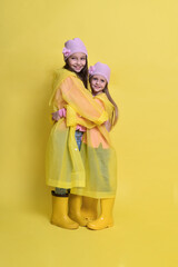 Two little sisters in yellow raincoats, boots and hats on a yellow background in autumn