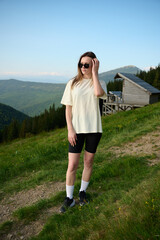 Obraz premium Woman walks on grassy mountain trail under warm sun, with wooden cabins and distant hills in background. Peaceful outdoor travel and nature lifestyle.