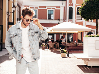 Portrait of handsome confident stylish hipster lambersexual model. Sexy man dressed in jeans jacket. Fashion male posing in the street. In sunglasses in Europe city. Outdoors at sunset
