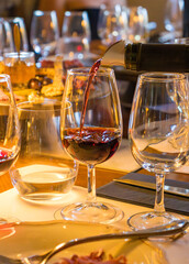 Close-up of a red wine glass on a dinner table with gourmet snacks, cheese, charcuterie, and appetizers. Warm evening atmosphere with elegant lighting, people in background, and luxury dining setting