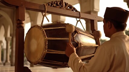 Musician in traditional attire strikes a large drum with mallets, showcasing rhythmic motion and cultural expression in a beautifully lit architectural setting