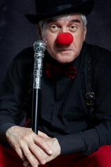 Dramatic portrait of a sad clown against a dark background with walking stick in hands.