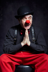 Clown on dark background with palms clasped together in a prayer gesture. Red clown nose and bowler hat on head.