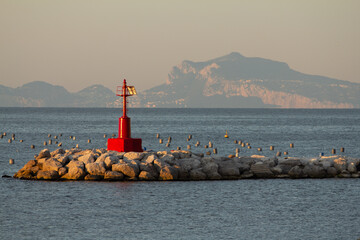 A striking red lighthouse stands on a rocky breakwater at sunset, with a misty mountain range in the background. Excellent for travel brochures, sailing magazines, and atmospheric coastal-themed conte