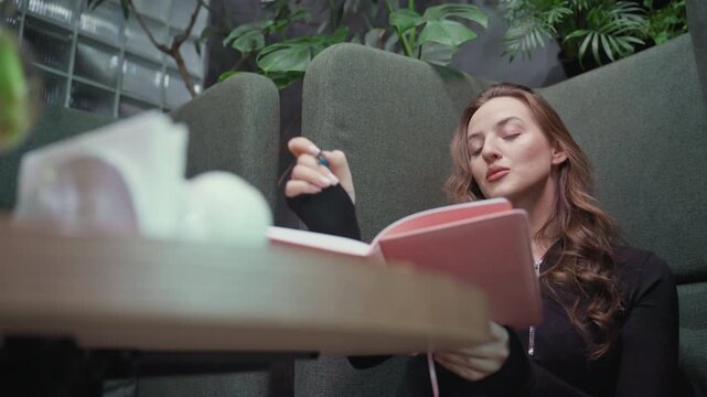 Blonde woman wearing black outfit sits with legs crossed in green booth, holding open pink notebook on lap and thinking deeply, surrounded by indoor plants and blurred tabletop decoration