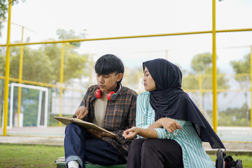 Two students studying outdoors together, discussing a tablet. A young man with headphones points at the screen, while a woman in a hijab attentively looks on. Collaboration and learning concept
