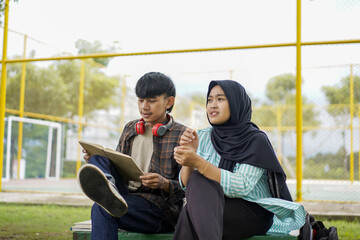 Two diverse young Asian students collaborating on a creative project outdoors, the boy holding a tablet and the girl in a hijab discussing ideas on a sunny day at their university campus