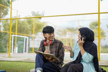 Two cheerful Asian university students sitting together outdoors on campus, a young man with a notebook and a woman in a hijab on a phone call