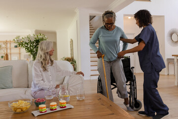 Senior woman using walking aid with caregiver while friend watches in living room
