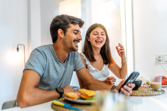 Happy couple laughing and using smartphone during breakfast at home - Powered by Adobe