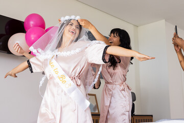 Bride-to-be celebrating with friends, wearing veil and sash, feeling joyful at home