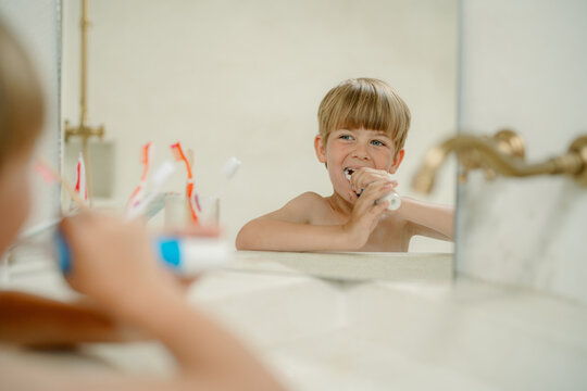Young boy enjoys brushing his teeth in a bright bathroom, looking at his reflection in the mirror. The ambiance is light and cheerful, highlighting a daily morning routine.