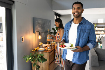 African American man carrying snacks while woman prepares drinks at home gathering