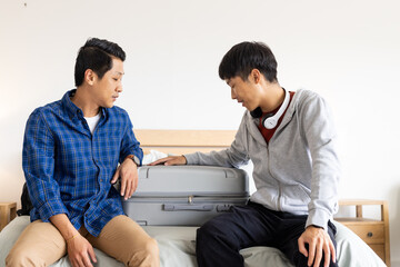Two men sitting on bed packing suitcase, preparing for travel together
